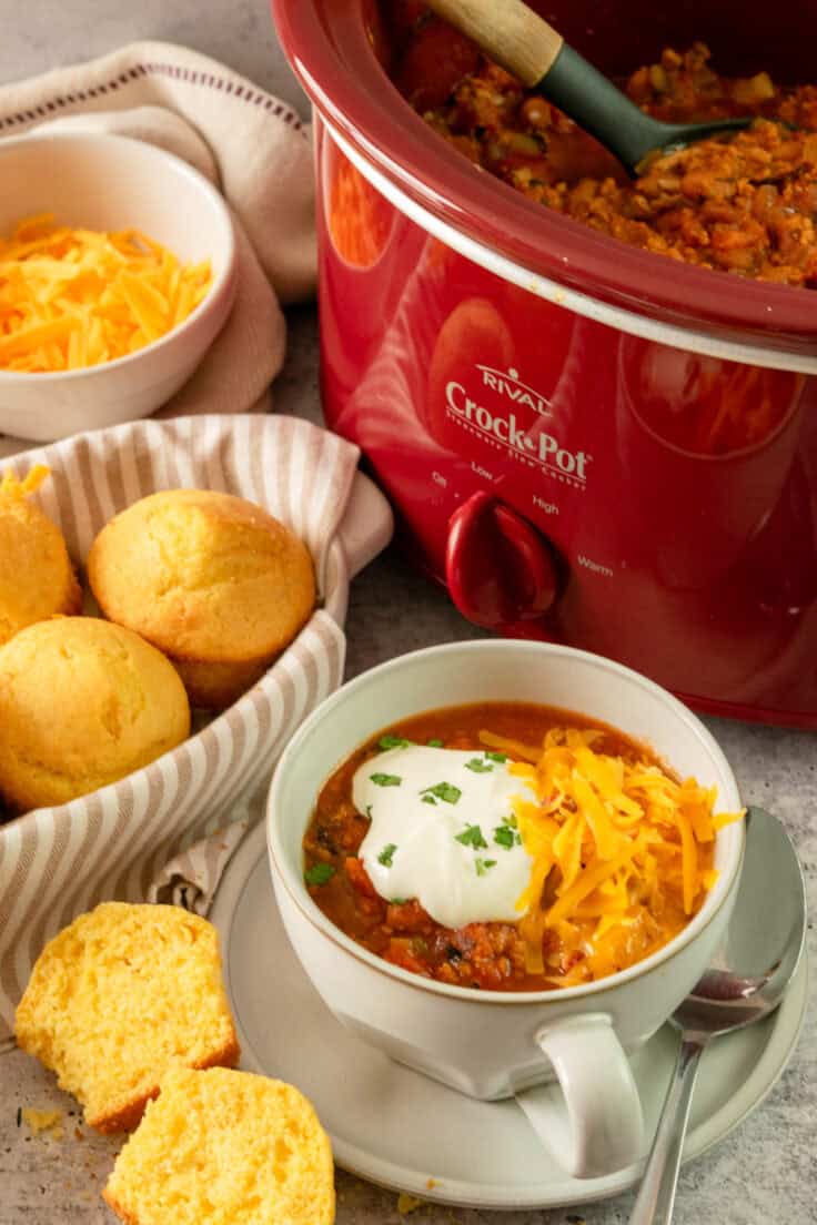 A red Crock Pot full of turkey chili, next to a bowl of of grated cheese, basket of cornbread muffins and a bowl of turkey chili with a spoon in it.