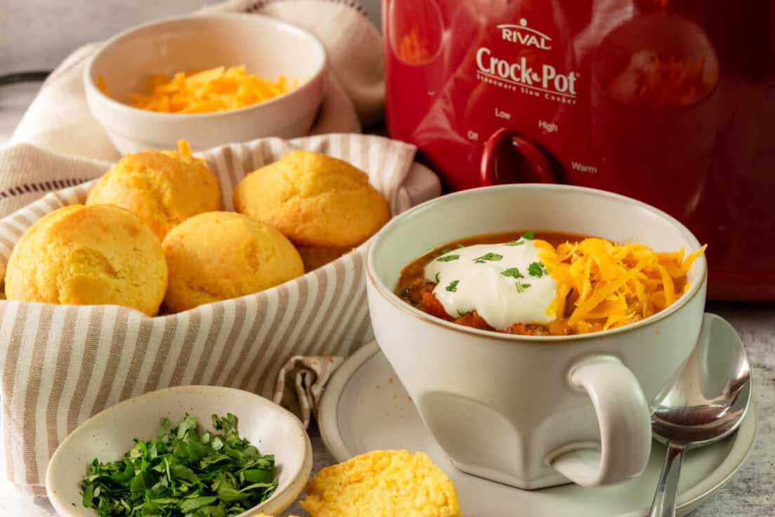 Bowl of turkey chili, topped with cheese and sour cream, next to a basket of cornbread and a red Crock Pot full of chili.