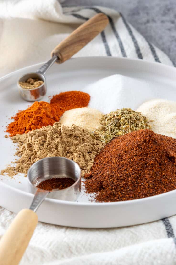 A close up view of chili powder, cumin, oregano, cayenne pepper and other spices piled in a bowl, next to two measuring spoons.