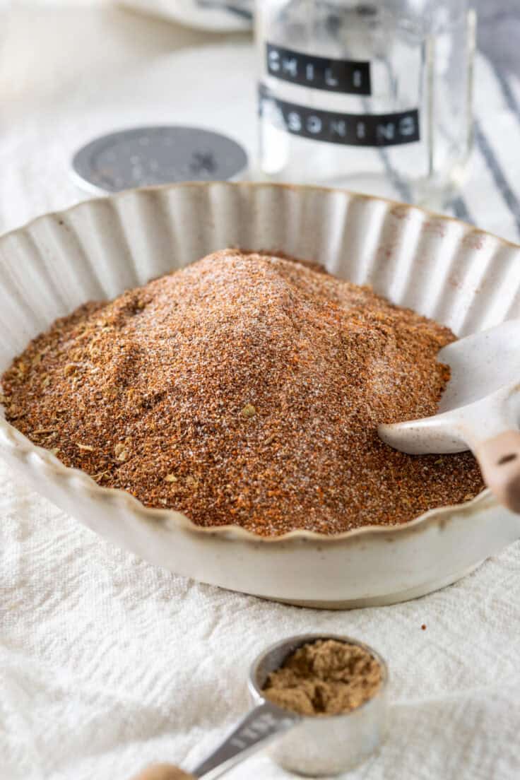 Homemade chili mix stirred together in a small bowl which is in front of an empty mason jar that has a label on it that says, chili seasoning.