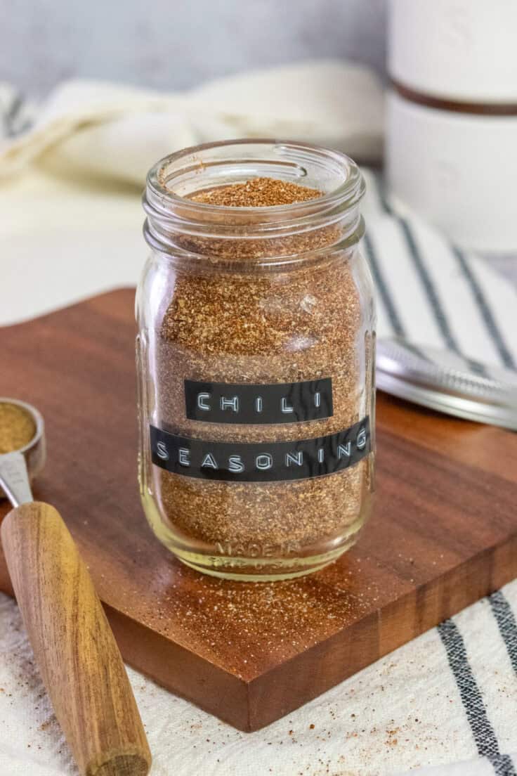 A mason jar with a black label on it that says, chili seasoning that's full of the spice mixture and next to a measuring spoon that has some cumin in it.