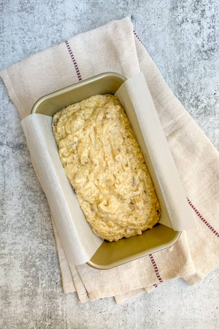 Banana bread batter spread into a loaf pan that’s lined with parchment paper.