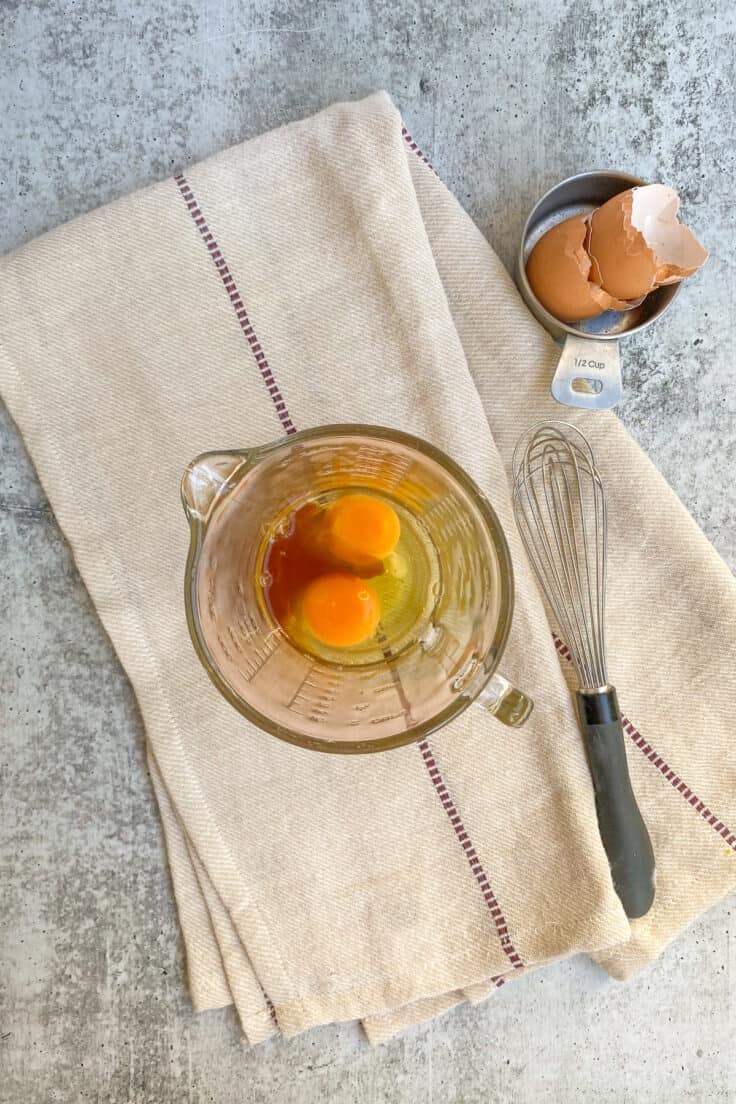 Two eggs and vanilla extract in a glass liquid measuring cup, next to whisk and cracked egg shells.