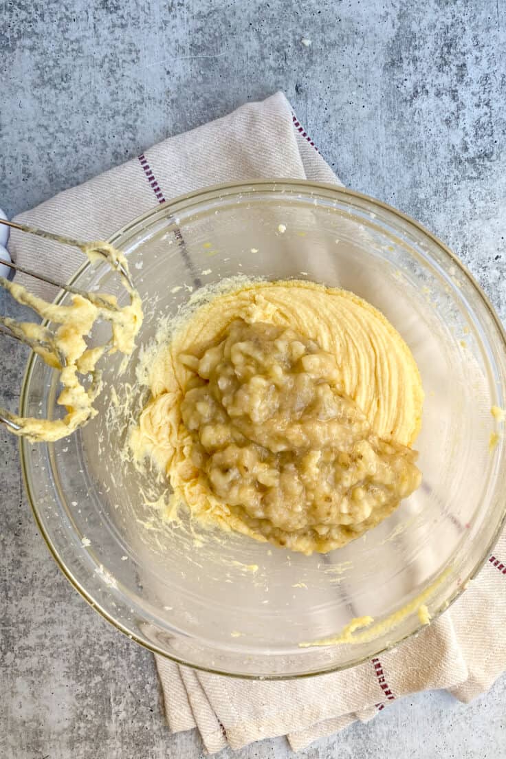 Mashed banana added to bowl of light and fluffy butter and sugar mixture.