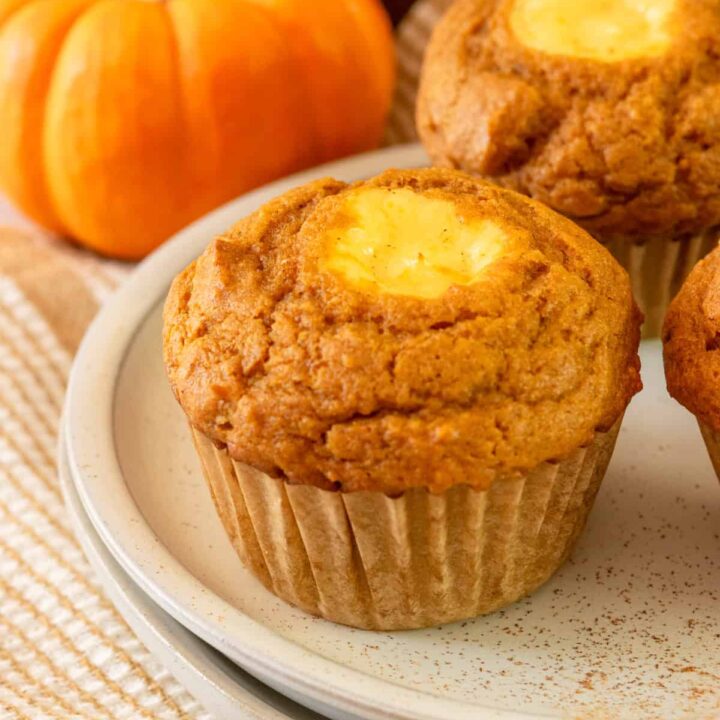 Homemade pumpkin cream cheese muffin on a plate in front of a mini pumpkin and more muffins.