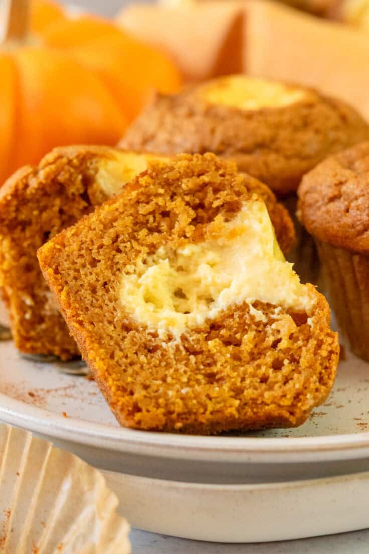 A homemade pumpkin muffin cut in half on a plate with other pumpkin muffins, showing the cream cheese center.