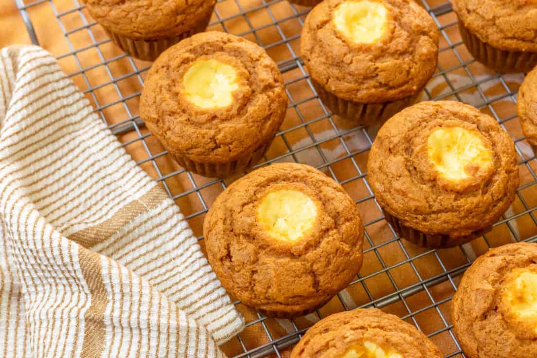 Homemade pumpkin muffins filled with cream cheese cooling on a rack with a tan and white towel next to it.