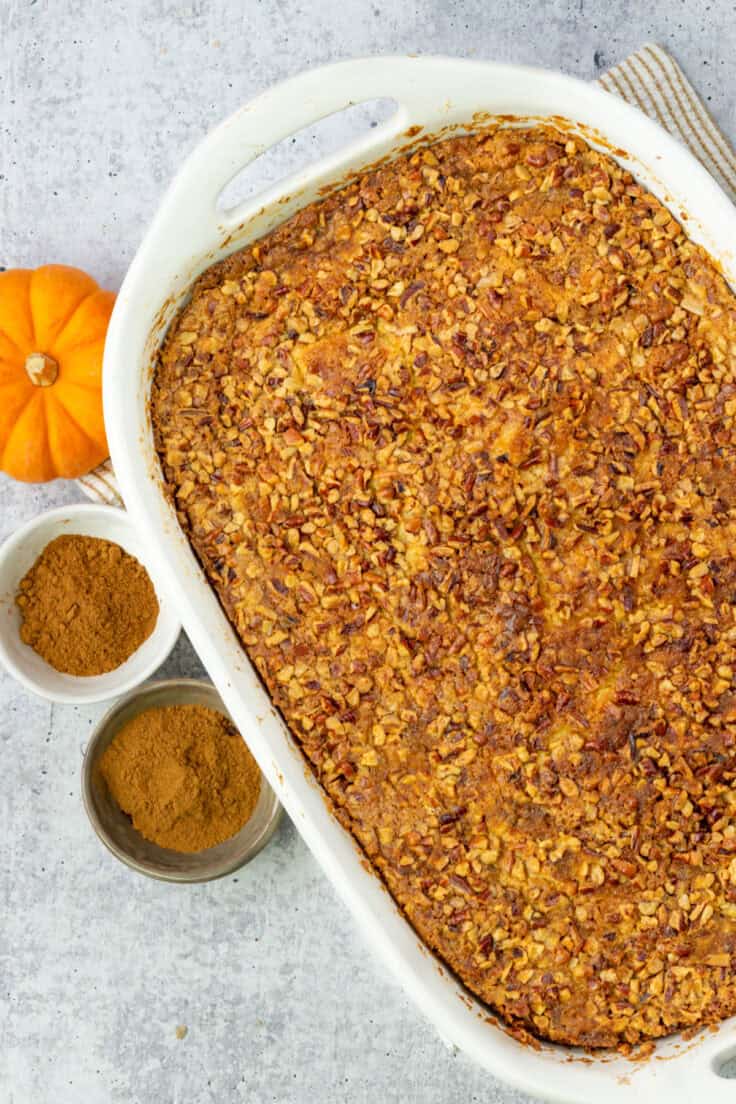 An overhead picture showing a pumpkin dump cake in a baking pan just out of the oven before it's been cut into. The dessert is next to a mini pumpkin and small bowls with cinnamon and pumpkin pie spice.