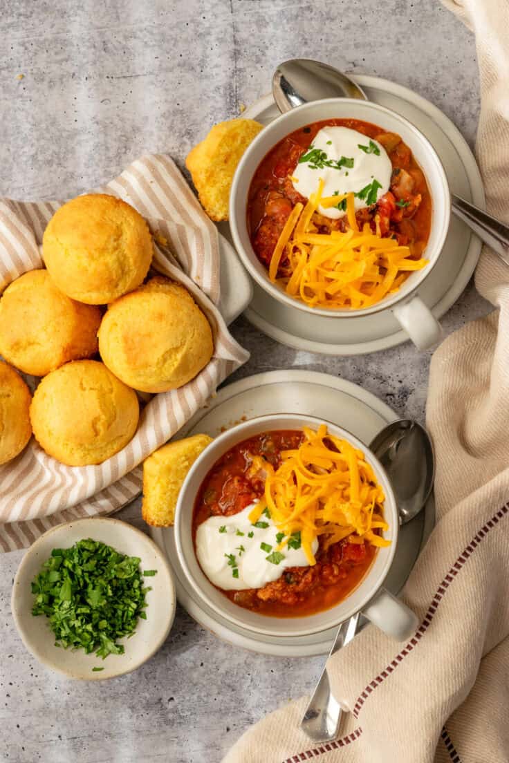 An overhead picture of two bowls of turkey chili topped with grated cheddar cheese, sour cream and cilantro. The bowls of chili are next to a basked of cornbread muffins and a small bowl of chopped cilantro.