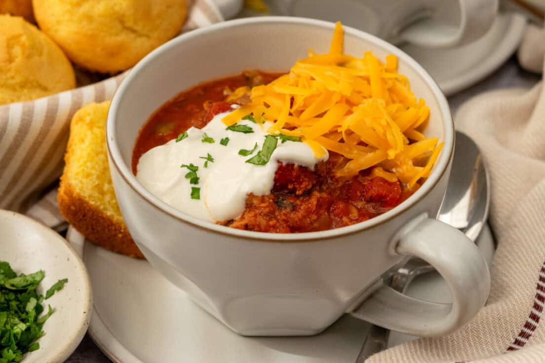 Close up view of a bowl of turkey chili topped with sour cream and cheese, on a saucer with a spoon and cornbread muffin on it.