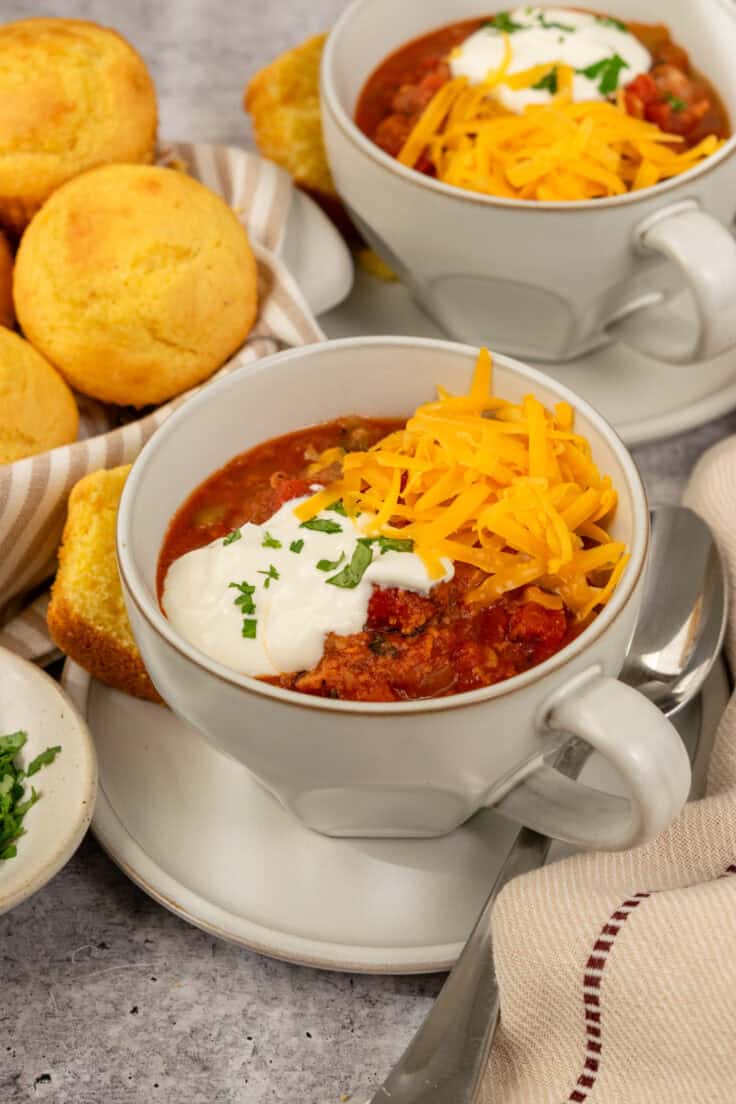 Two bowls of turkey chili made in slow cooker, topped with sour cream and cheese with cornbread muffins on the side.