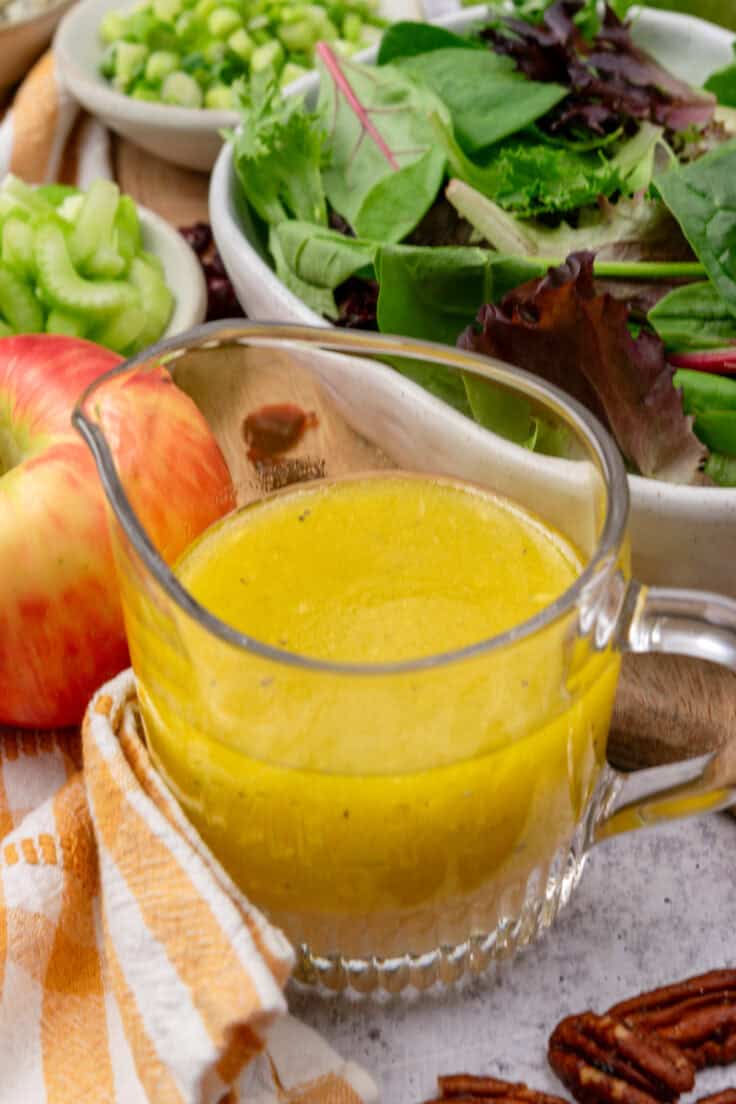 A glass jar of homemade apple cider maple vinaigrette sitting next to an apple and bowl of salad greens.