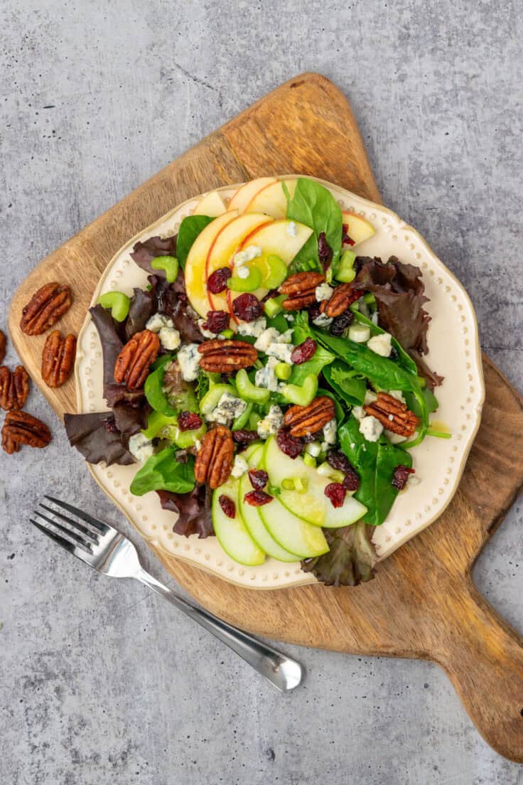 Apple pecan salad with crunchy pecans and chewy dried cranberries on top, drizzled with vinaigrette, on a small salad plate which is sitting on a wooden board with a fork next to it.