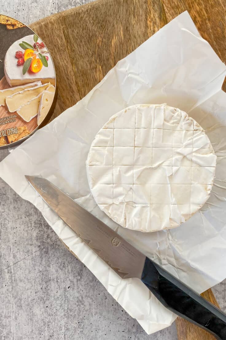 A wheel of brie cheese laying on its wrapper with a knife on it, showing how the cheese has been scored on top before baking.