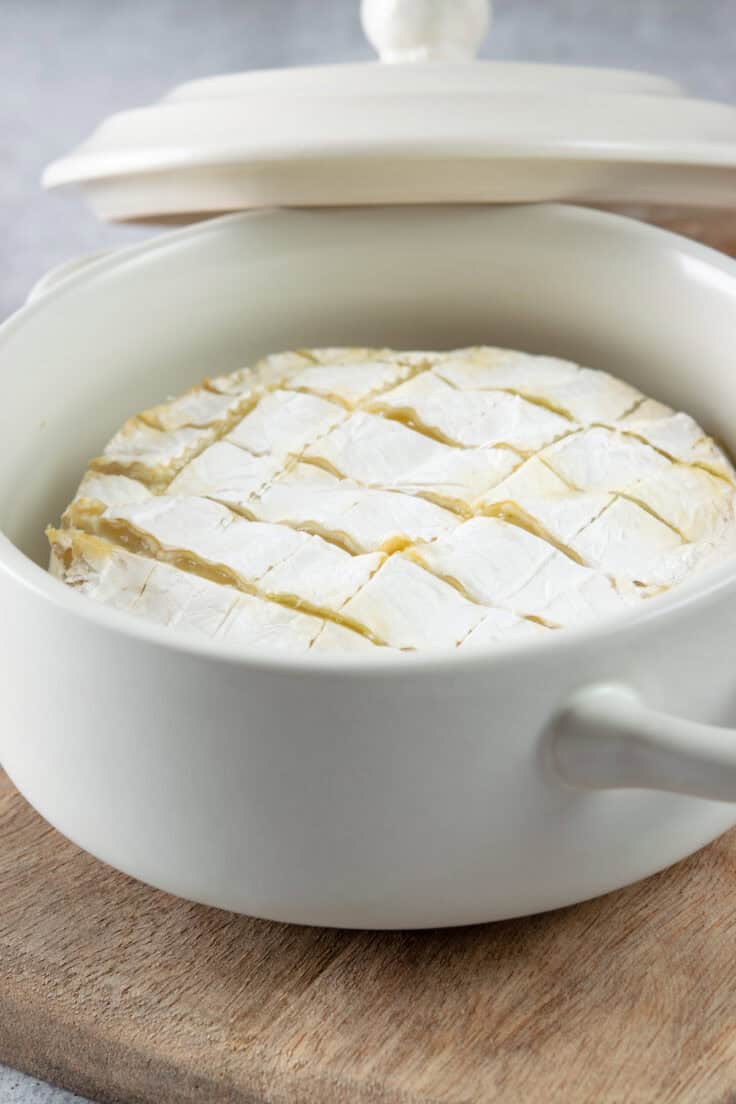 Brie cheese that's been scored in a crosshatch pattern on top, in a brie baker with a lid next to it, showing the melted cheese.