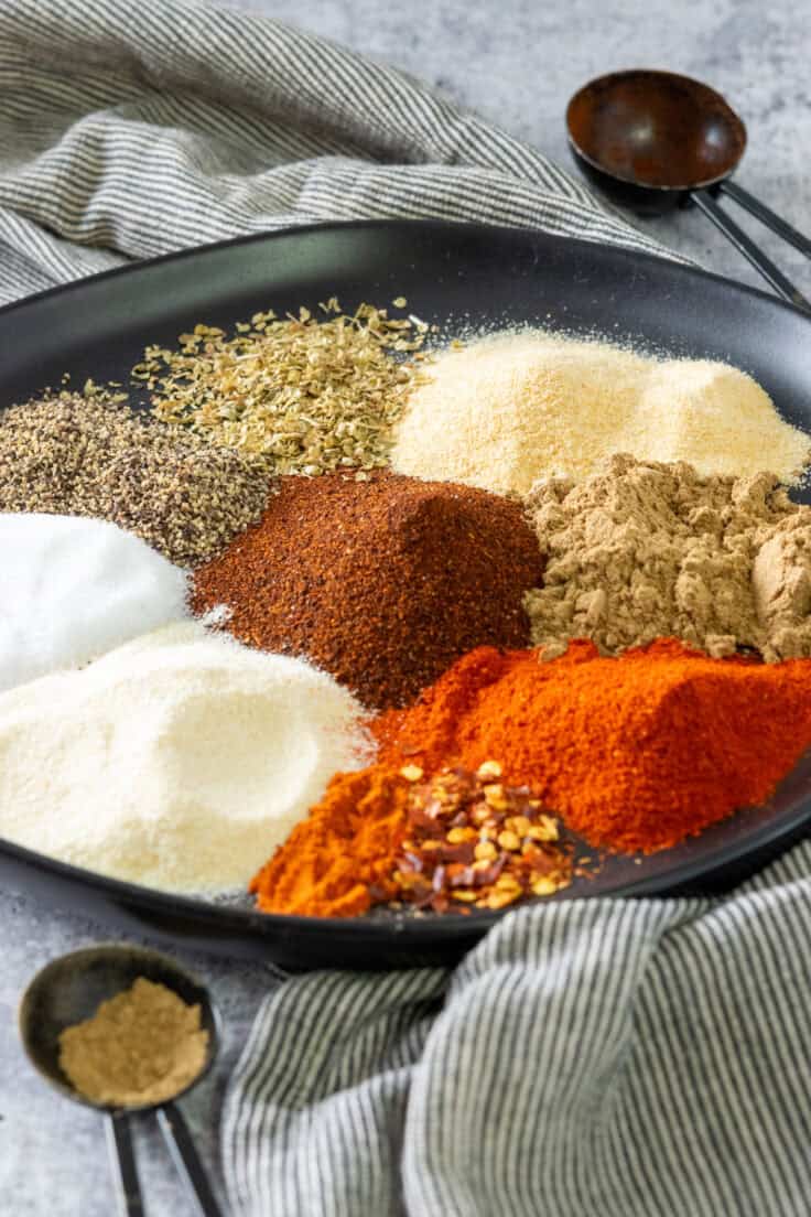 A plate with measured out spices to make a taco season blend, including chili powder, cumin, garlic powder, onion powder, oregano, smoked paprika, cayenne pepper, red pepper flakes, and salt and pepper.