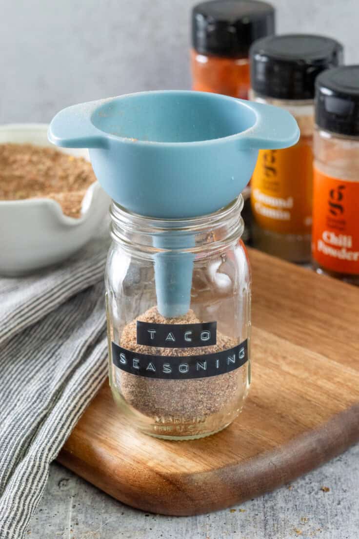 A small funnel in a spice jar that's labeled taco seasoning, with the spices being poured into it.