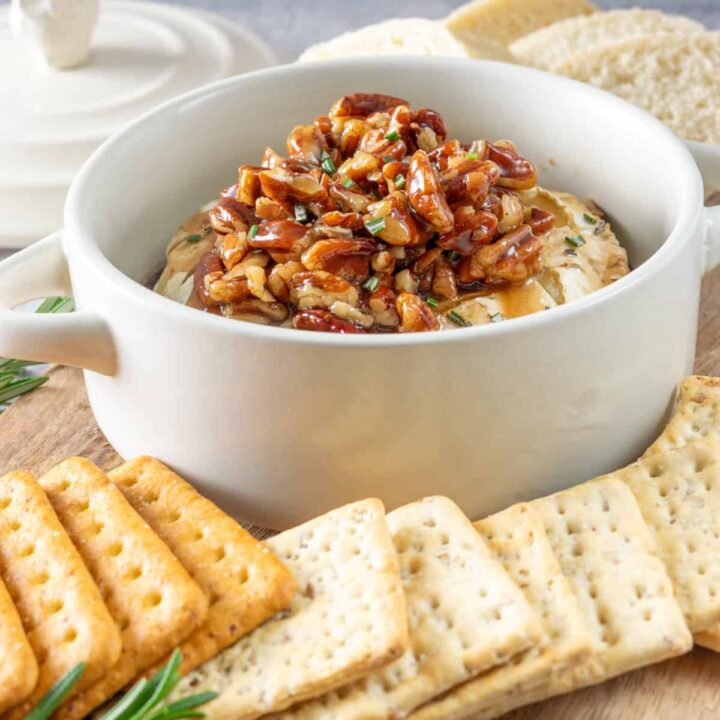 Baked brie in a brie baking dish topped with maple pecans, all on a board with crackers and bread for dipping.