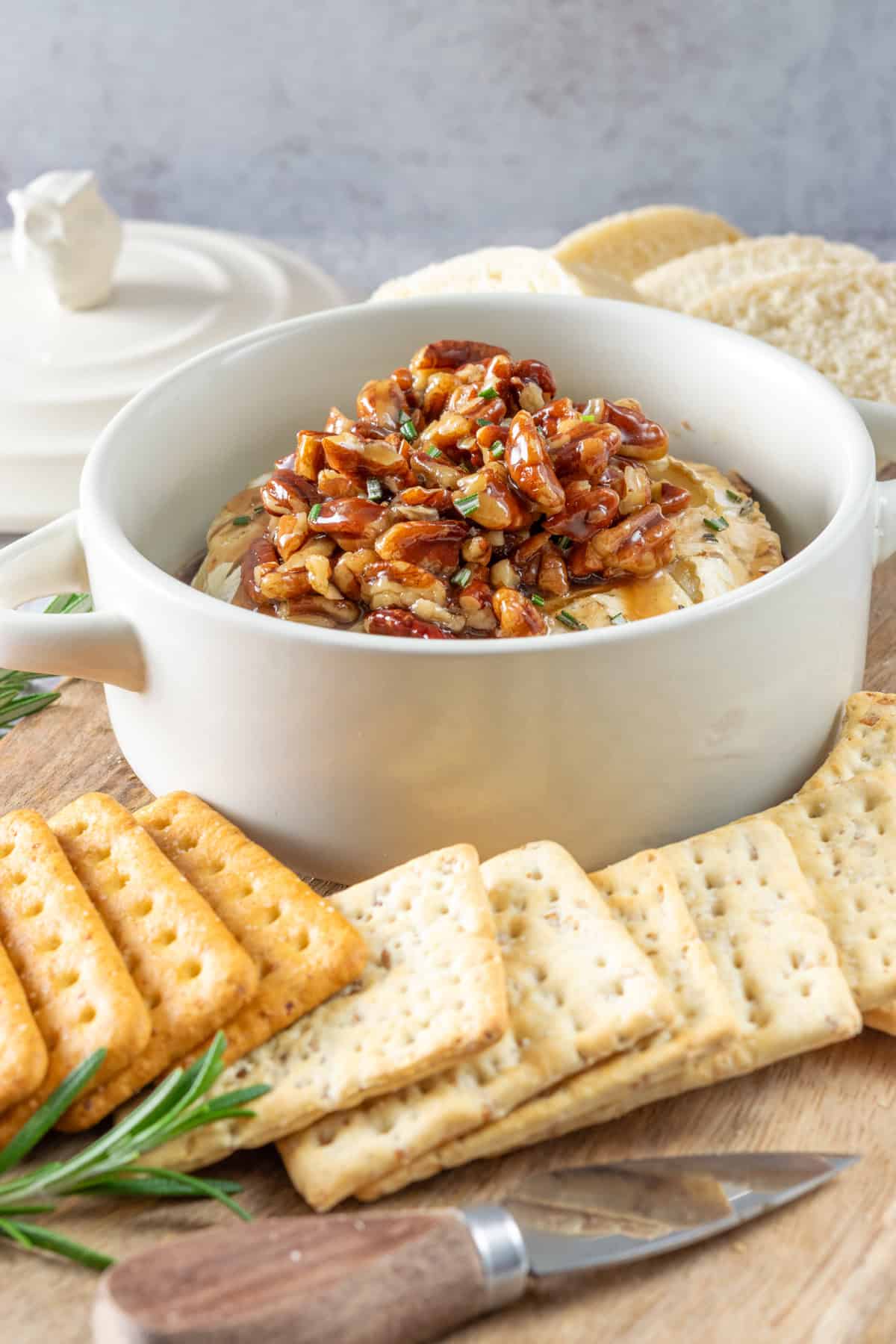 Baked brie in a brie baking dish topped with maple pecans, all on a board with crackers and bread for dipping.