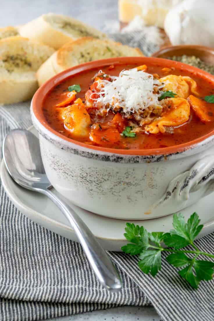 A warm and hearty bowl of sausage tortellini soup with parmesan cheese melting on top, that's garnished with Italian parsley and next to slices of toasted Italian bread and a block of parmesan cheese.