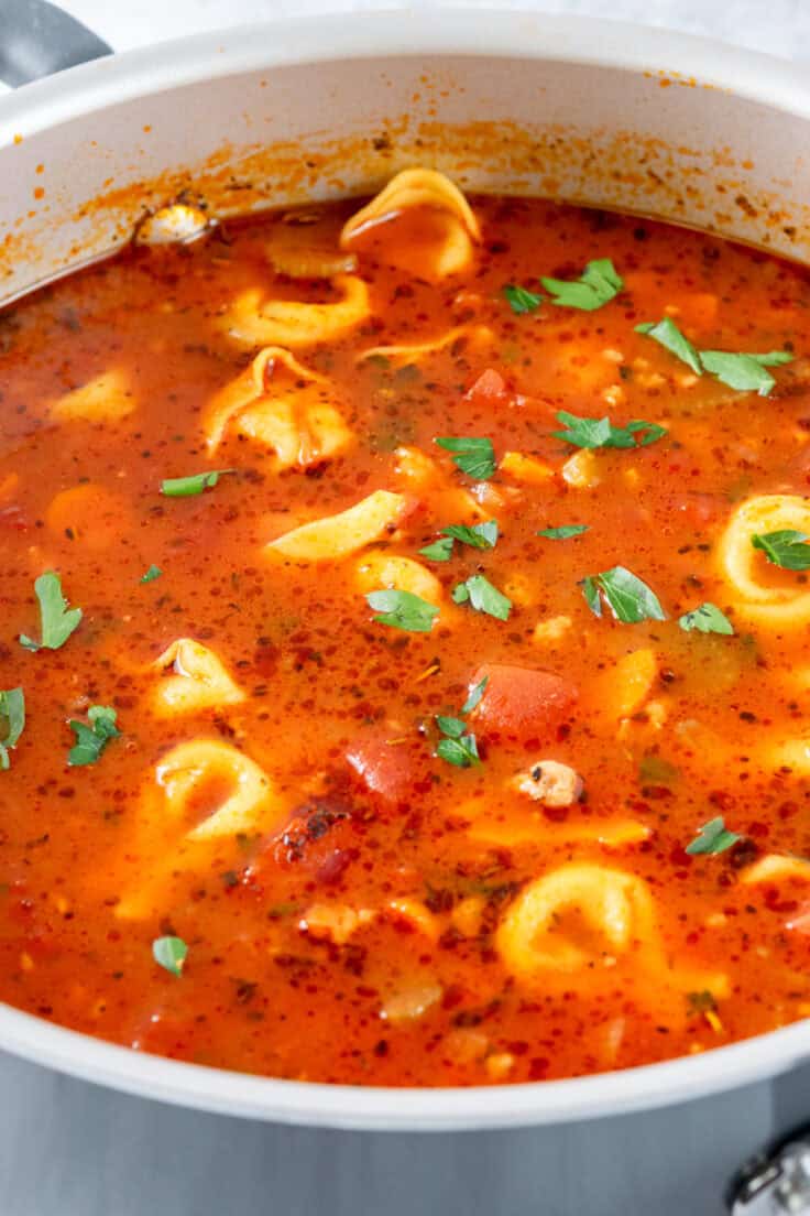 A stock pot full of hot bubbling sausage tortellini soup with bits of torn parsley on top.