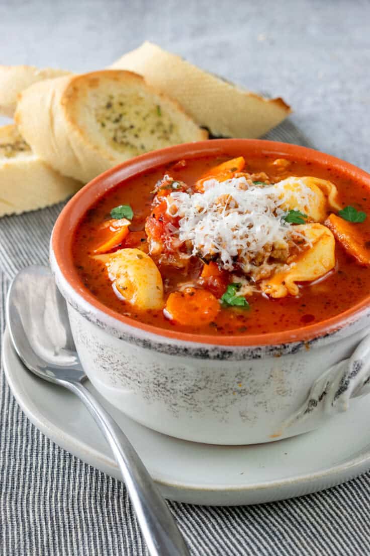 Bowl of sausage tortellini soup on a plate with a spoon. The soup is full of tortellini, carrots, lots of meat and has grated parmesan on top.