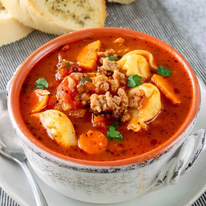 Bowl of sausage tortellini soup on a plate with a spoon and toasted baguette.