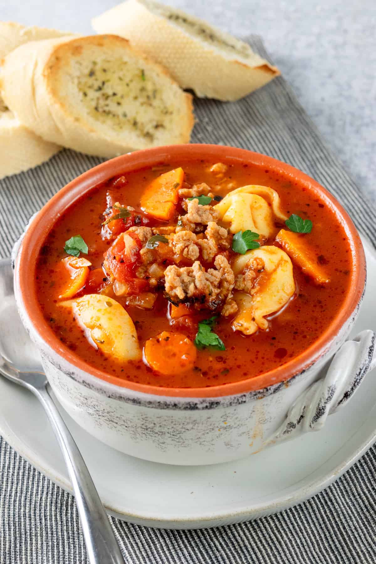 Bowl of sausage tortellini soup on a plate with a spoon and toasted baguette.