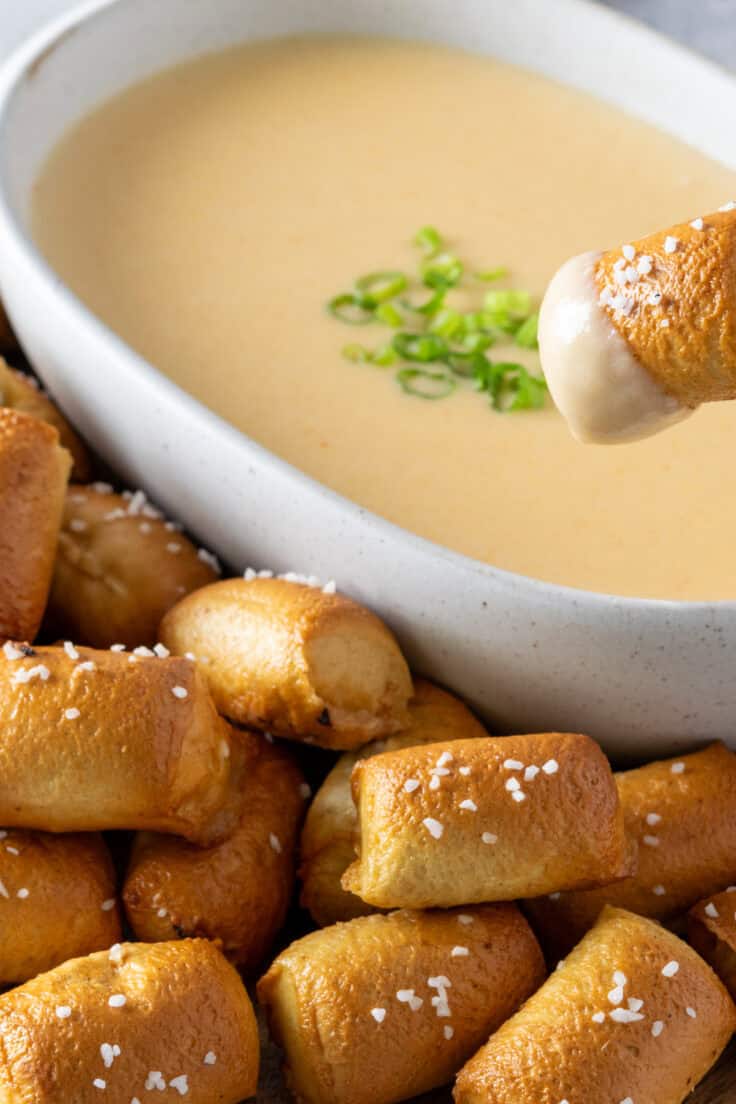 A salty pretzel bite that's being dipped in a bowl of beer cheese dipping sauce that's surrounded by pretzel bites on a tray.