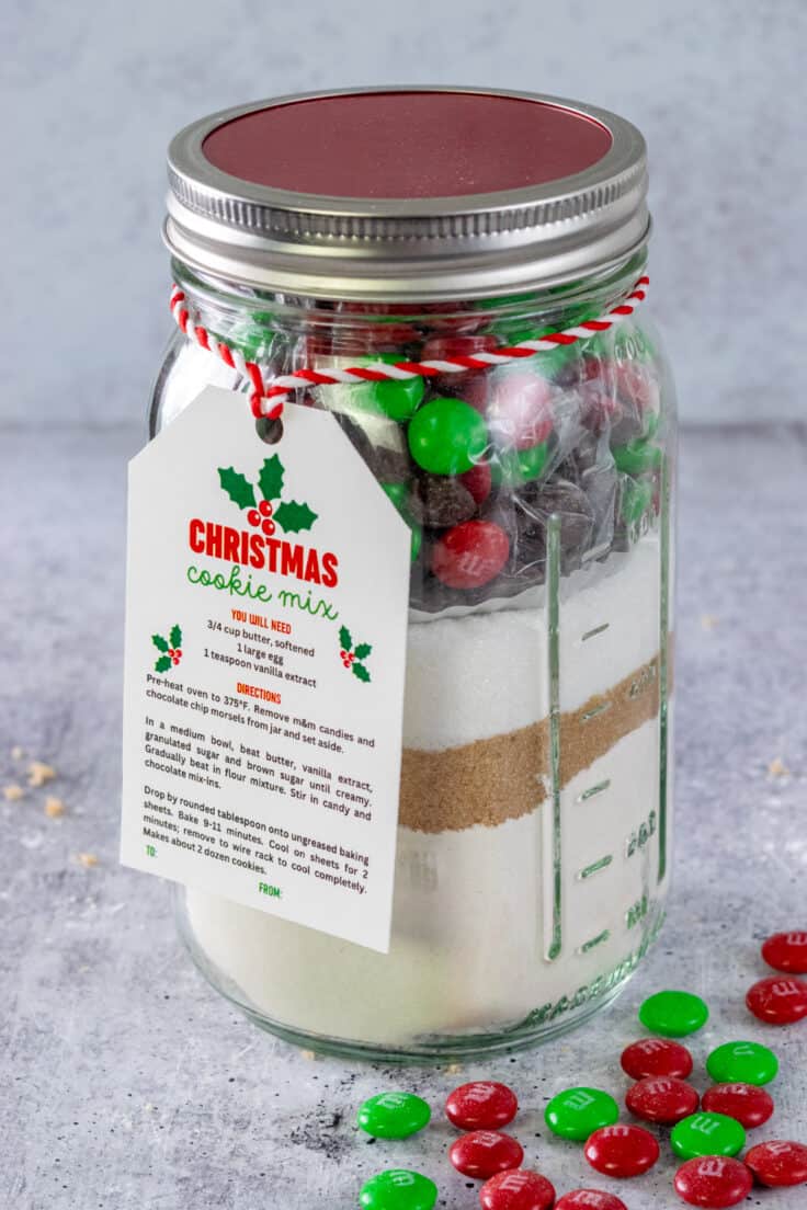 Homemade Christmas Cookie Mix layered in a mason jar that has a printable gift tag on it and some red and green holiday m&m's scattered around the jar.