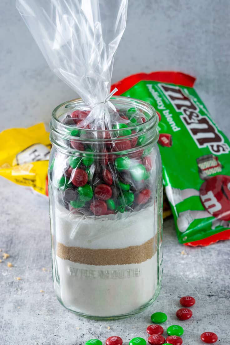 A bag filled with chocolate chips and red and green holiday m&m's placed inside the cookie mix in a jar.