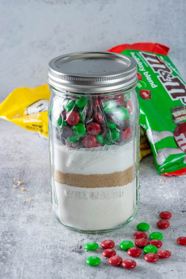 Homemade Christmas cookie mix in a jar sitting next to bags of Nestle chocolate chips and holiday m&m's with a few red and green m&m's scattered around the jar.