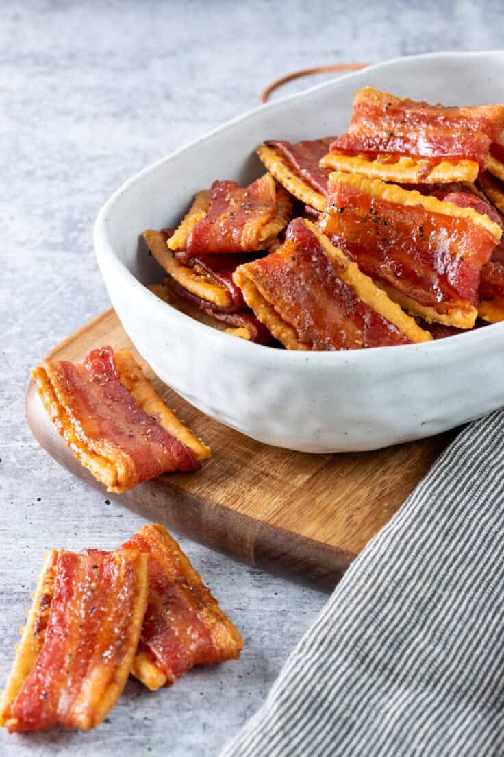 Candied bacon crackers in a small serving dish which is sitting on a board and next to a ticking-stripe towel. There are three mouth-watering bacon crackers outside of the dish.