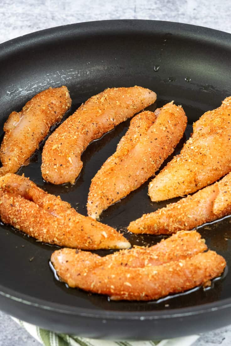 Seasoned chicken tenderloins placed in a skillet with olive oil with the raw side of the chicken facing up.