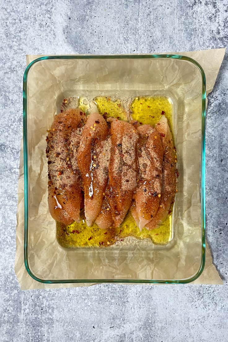 Olive oil poured over seasonings and raw chicken tenderloins that are in a glass dish.