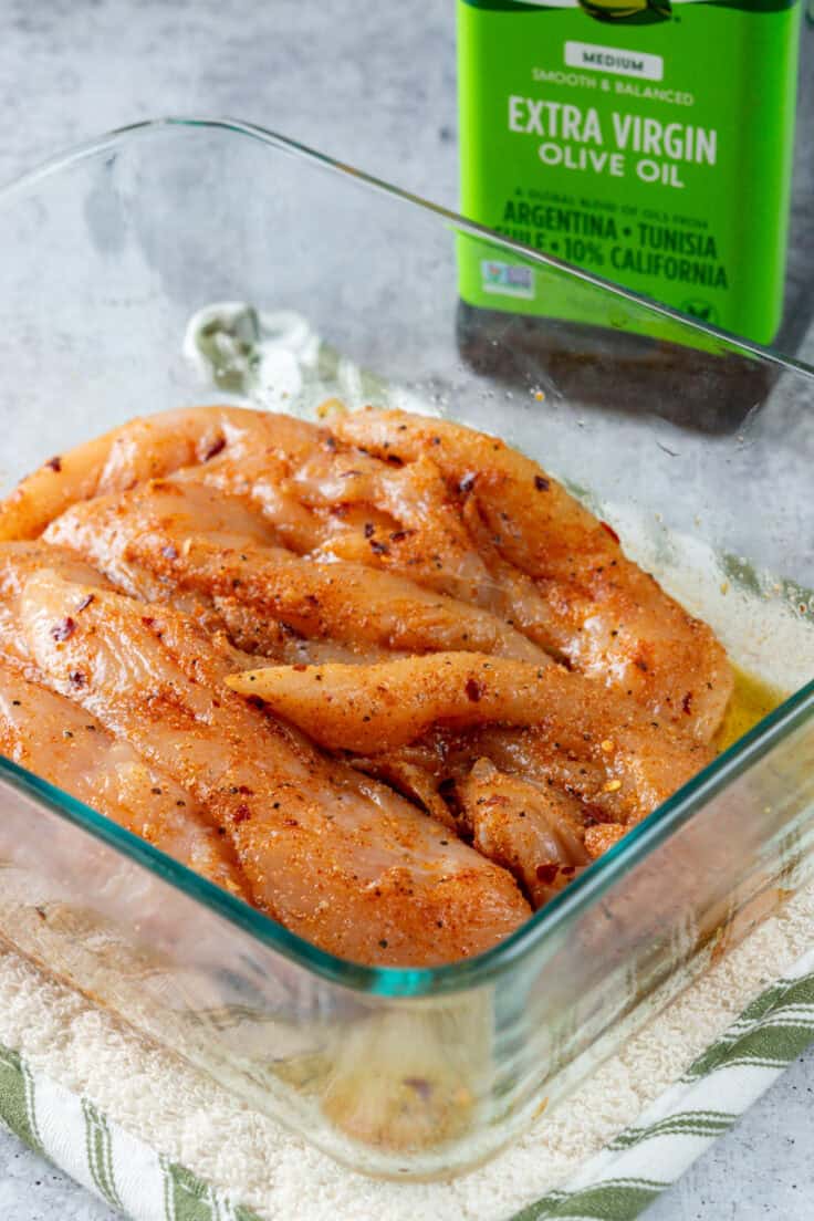 A glass dish containing raw chicken tenderloins that are fully coated in seasoning and olive oil. The dish is next to a bottle of extra virgin olive oil.