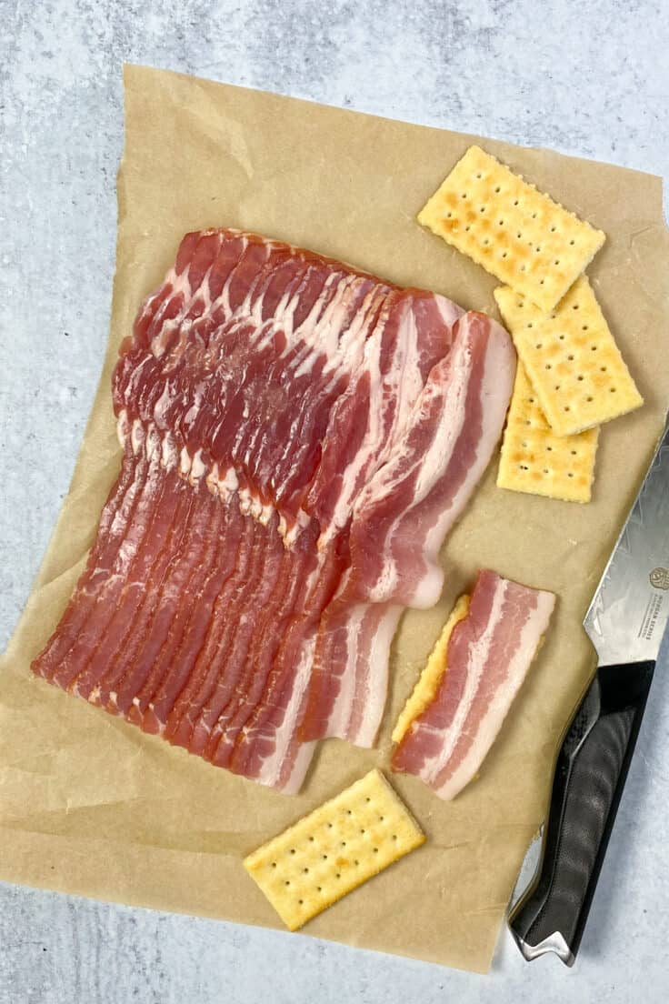 Bacon strips on a piece of parchment paper next to some club crackers and a knife. A strip of bacon is cut and laying on top of a club cracker to indicate the length the bacon should be cut for this recipe.