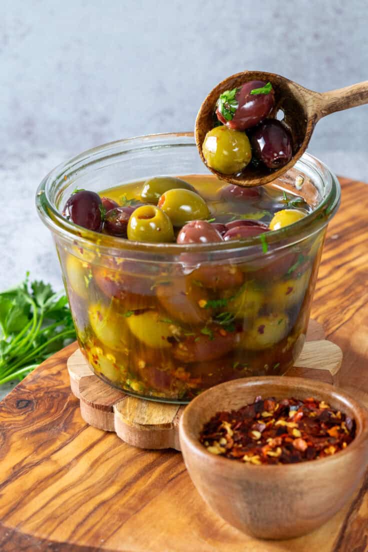 A glass bowl of marinated olives sitting on a wood board next to a small bowl of red pepper flakes. Three marinated olives are being lifted out of the bowl with a wooden olive spoon.