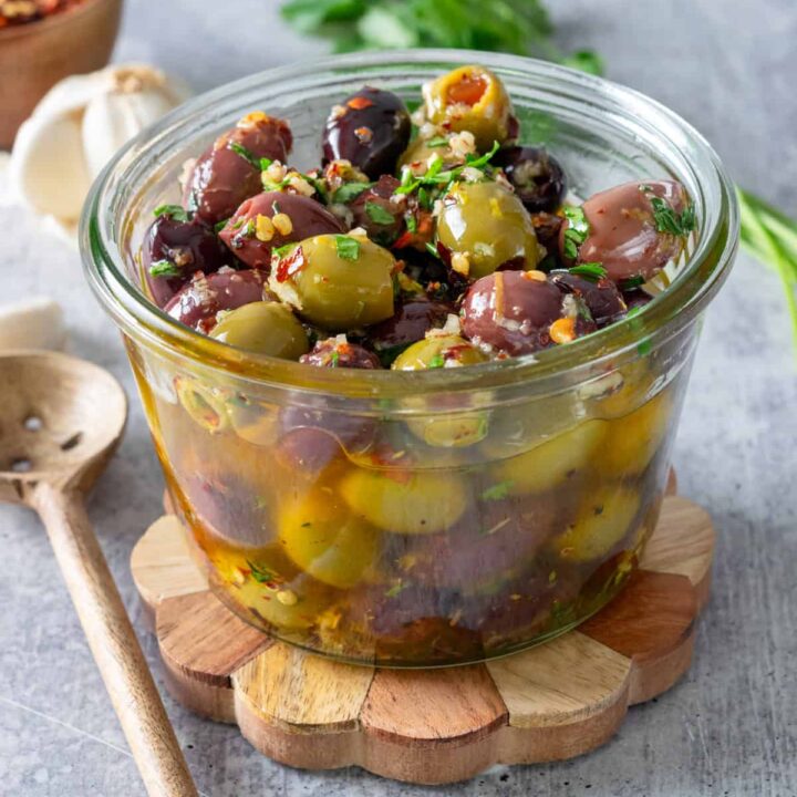 Homemade marinated olives in a glass bowl next to an olive spoon, garlic bulb and herbs.