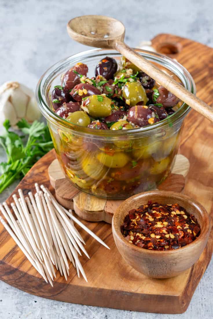 A bowl of homemade marinated olives with an olive spoon resting on top. The olives are on a wooden serving board next to some toothpicks, a small bowl of red pepper flakes, fresh herbs and garlic bulb.