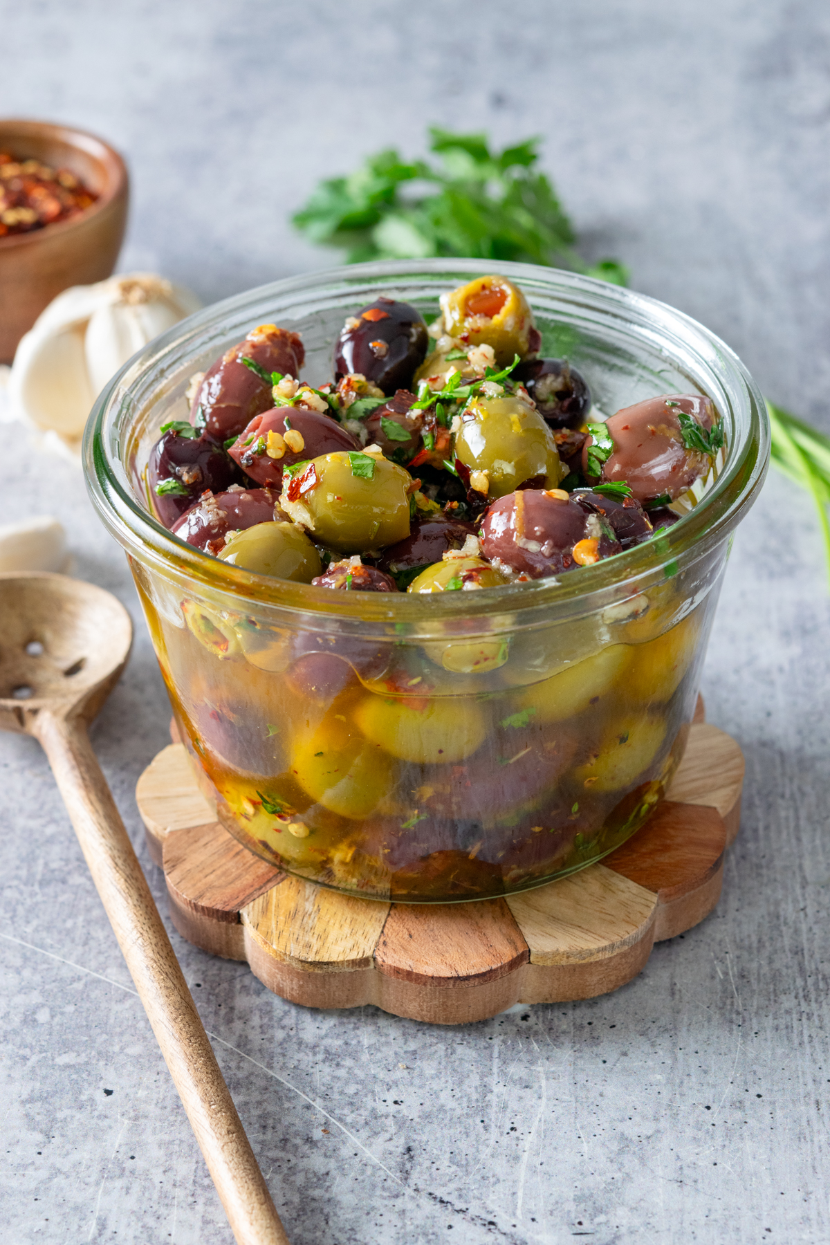 Homemade marinated olives in a glass bowl next to an olive spoon, garlic bulb and herbs.