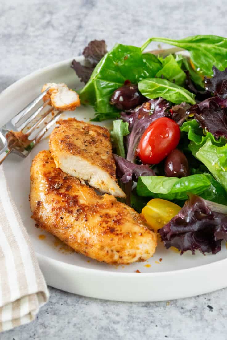 Chicken tenderloins on a plate with one that's been cut into with a piece of chicken on a fork. The chicken is plated with a healthy salad.