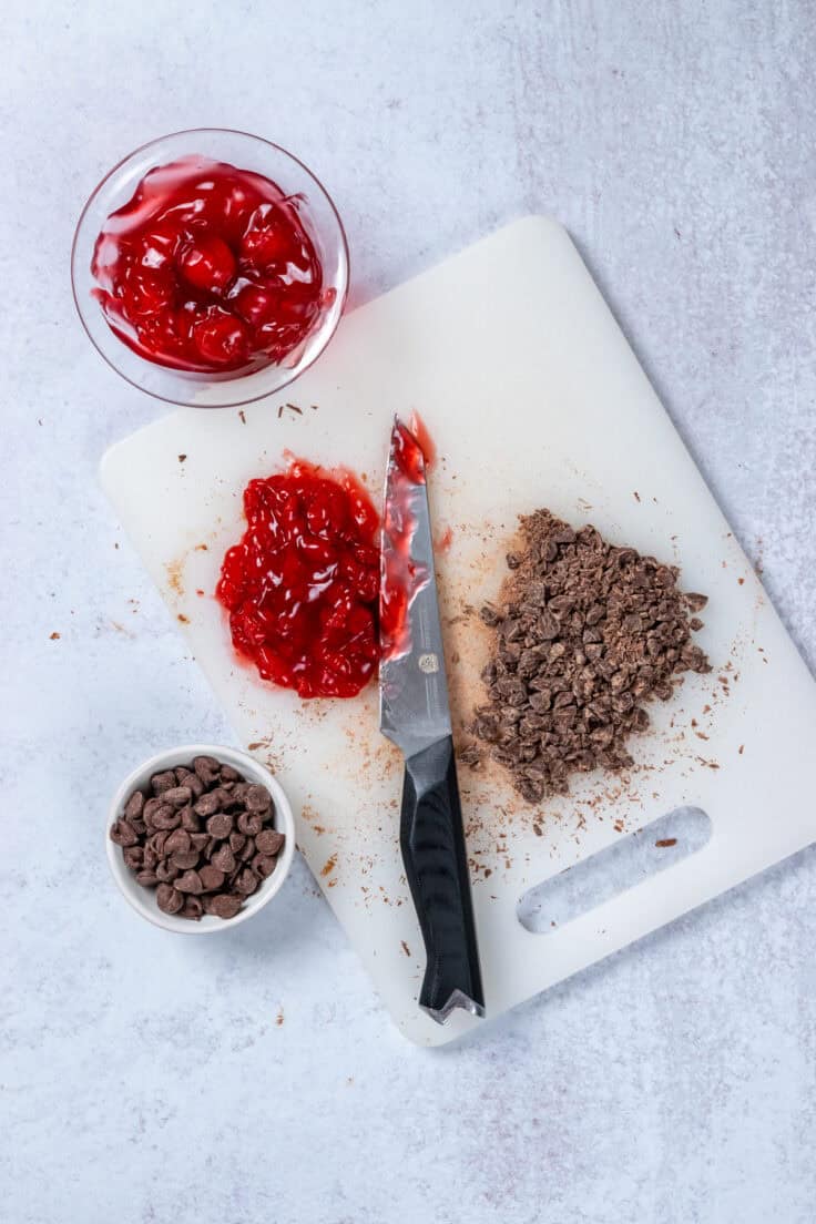 A cutting board with knife showing a pile of the cherries roughly chopped and the chocolate chips finely chopped.