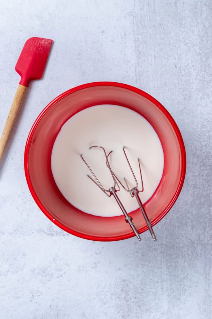 A red mixing bowl full of milk and sugar with two beaters in it and a red rubber spatula sitting next to it.