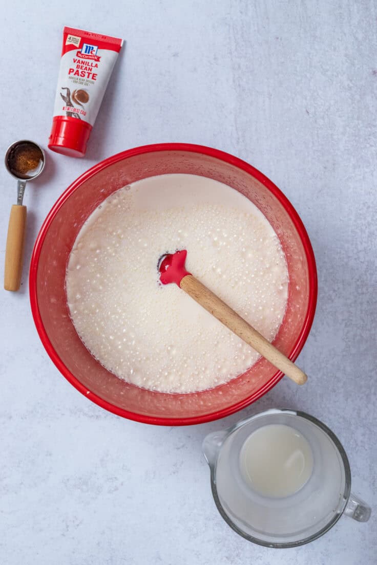 A red mixing bowl that contains all the base ice cream mix ingredients (heavy cream, milk, sugar, salt and vanilla) next to a tube of vanilla bean extract a measuring cup that had heavy cream in it.