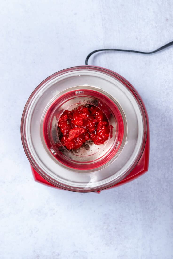 An overhead view of a red Cuisinart automatic ice cream maker with cherries and chocolate added on top.