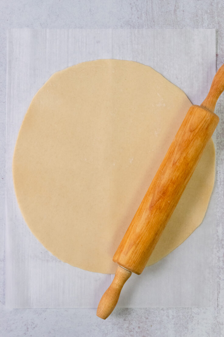 One Pillsbury pie crust on a piece of parchment paper being rolled thinner with a rolling pin.