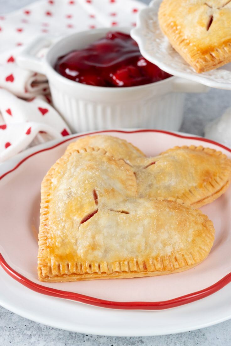 Two heart-shaped cherry hand pies on a pink plate next to a bowl of cherry pie filling and a platter with more cherry hand pies.