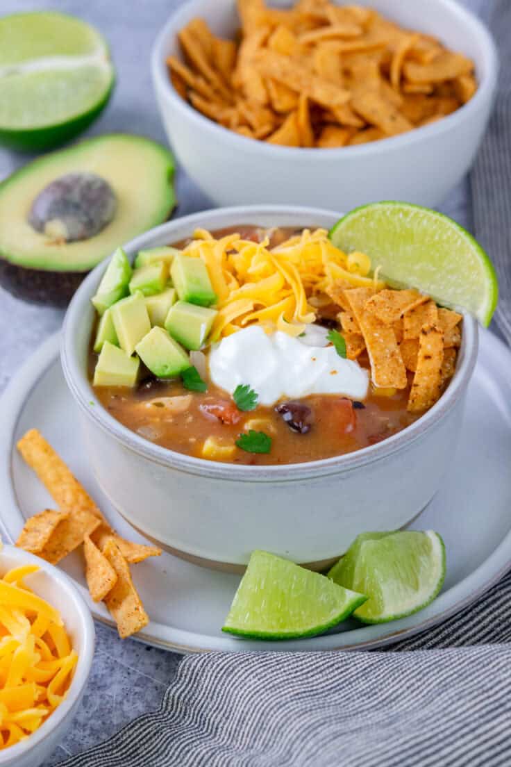 A bowl of creamy chicken tortilla soup garnished with avocado, sour cream, cheese, tortilla strips and lime wedge. The bowl of soup is next to half an avocado, half a lime, and a small bowl full of tortilla strips.