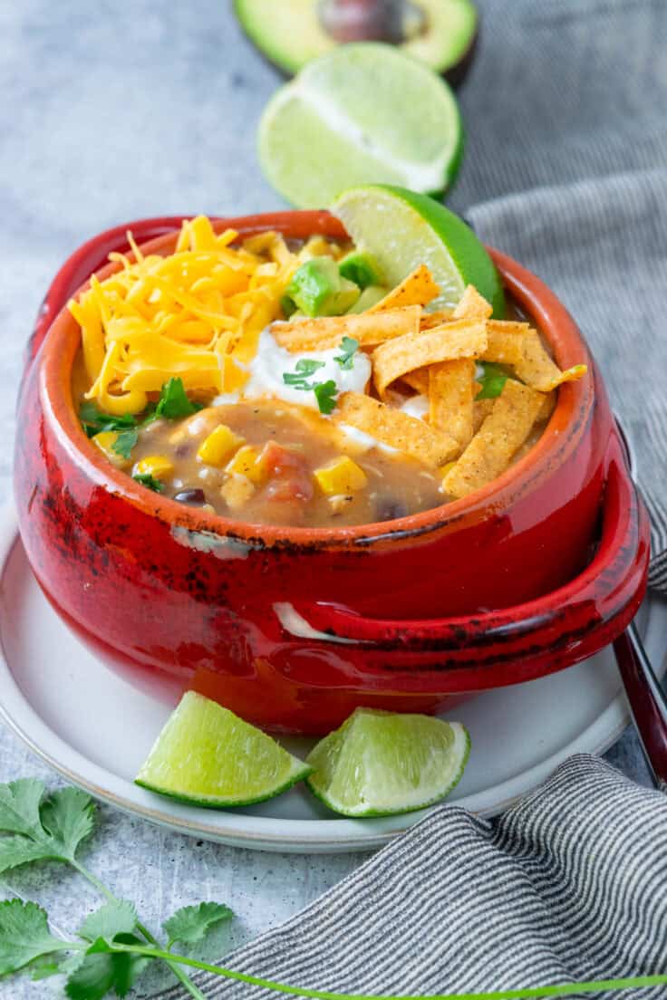 A bowl of homemade chicken tortilla soup with lots of corn in it, that's topped with cheese, sour cream, tortilla strips, cilantro and lime. The bowl is on a plate that has a spoon resting on it and two pieces of lime.